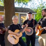 four musicians with mandolin, guitar, banjo, bouzouki outdoors smiling